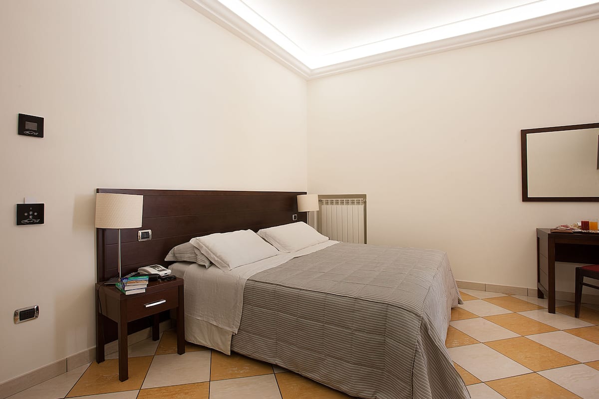 A hotel room with a bed, a nightstand with a lamp, a chair, and a mirror on the wall.