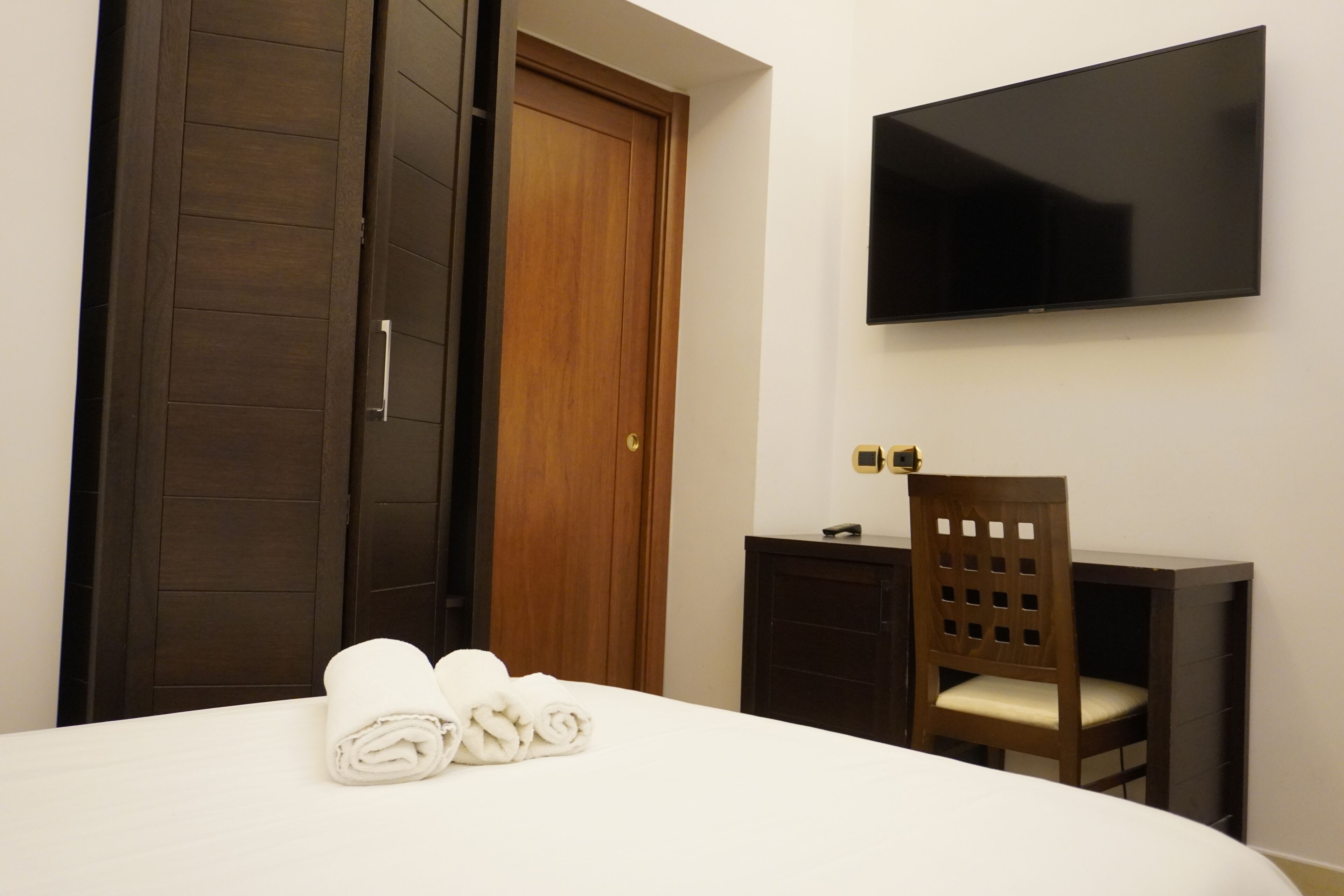 A hotel room with a bed, a TV mounted on the wall, a wooden wardrobe, and a desk with a chair.
