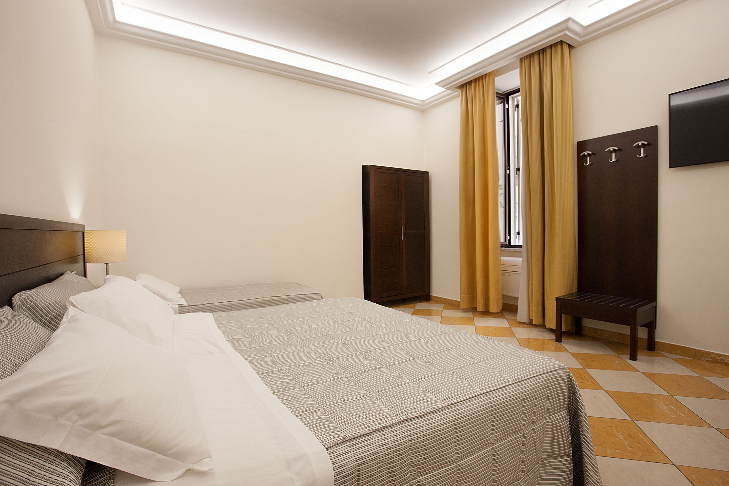A hotel room with two beds, a wardrobe, a bench, and a TV mounted on the wall.