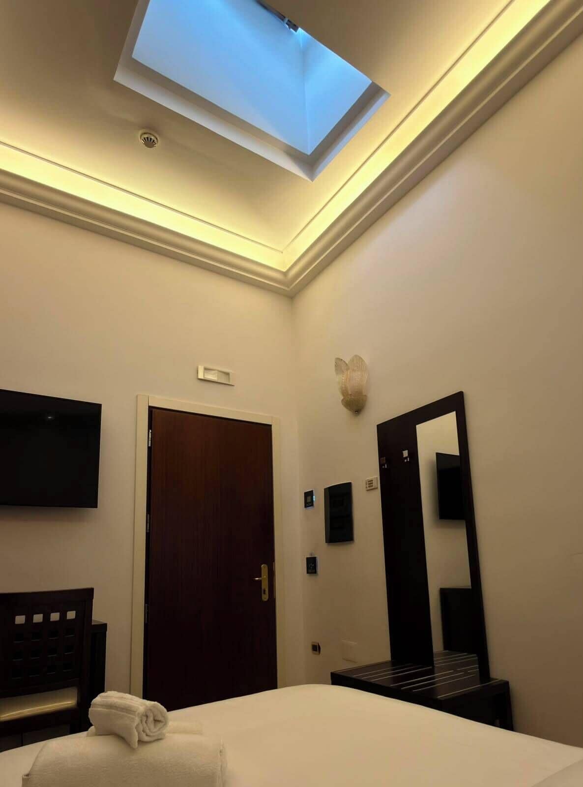 A hotel room with a skylight, a bed, a TV, a mirror, and a door.