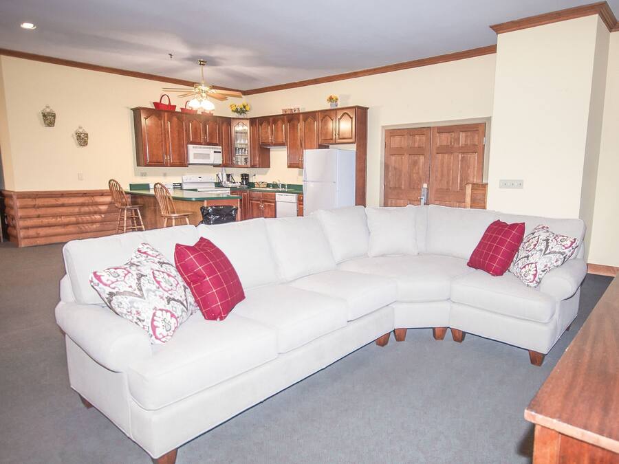 A living room with a white sectional sofa, wooden cabinets, and a kitchen area in the background.