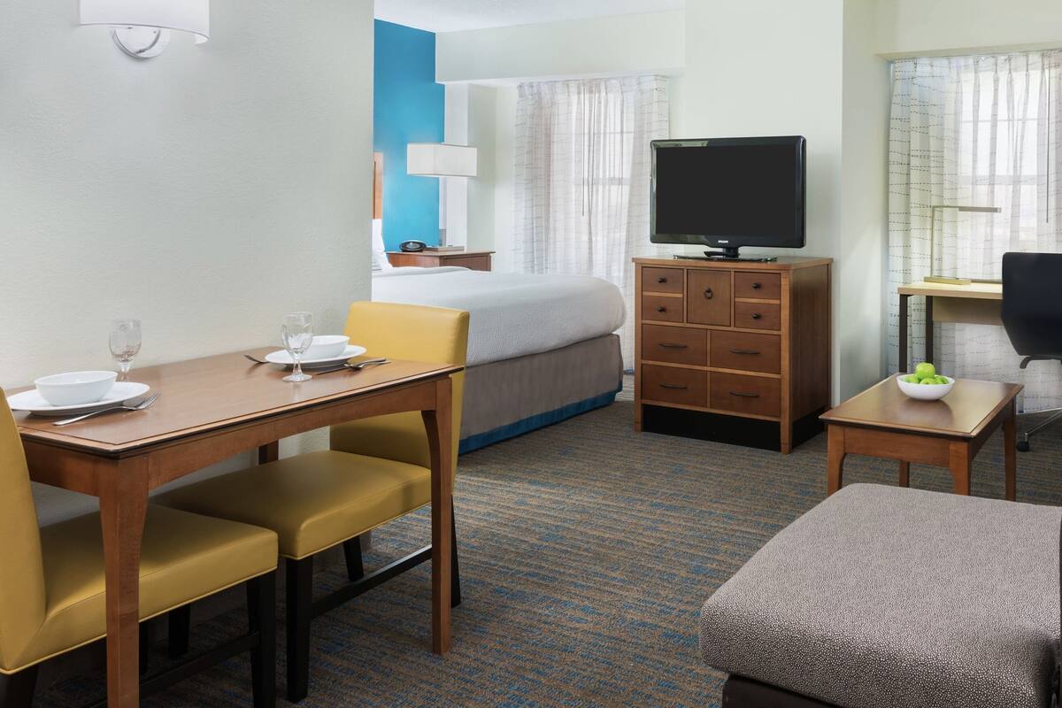 A hotel room with a bed, a desk with a computer, a TV, a dining table with chairs, and a small table with a bowl of fruit.