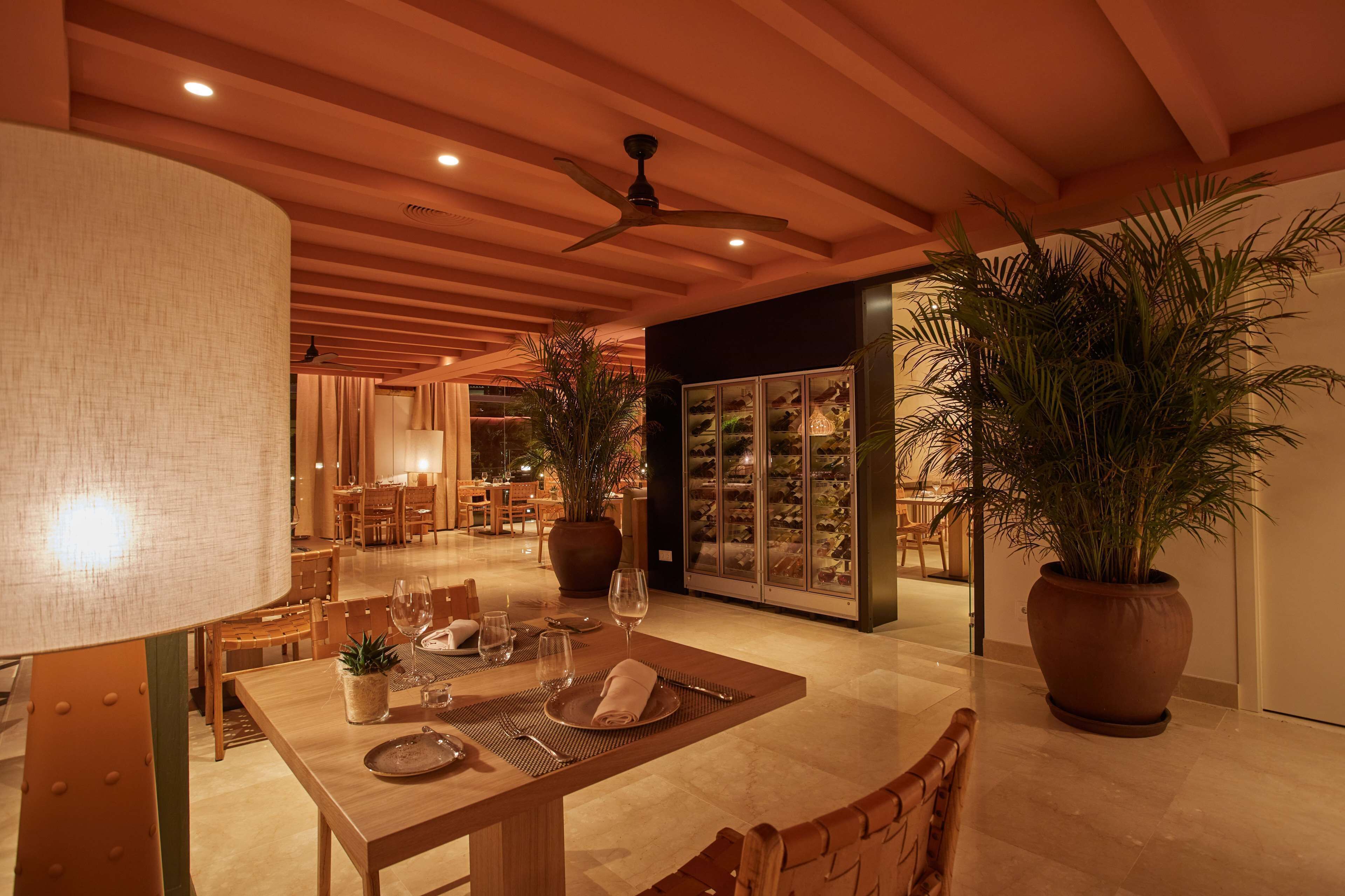 Un comedor bien iluminado con una mesa de madera dispuesta para dos personas, rodeada de sillas. Se observan plantas en macetas y un acuario grande al fondo.