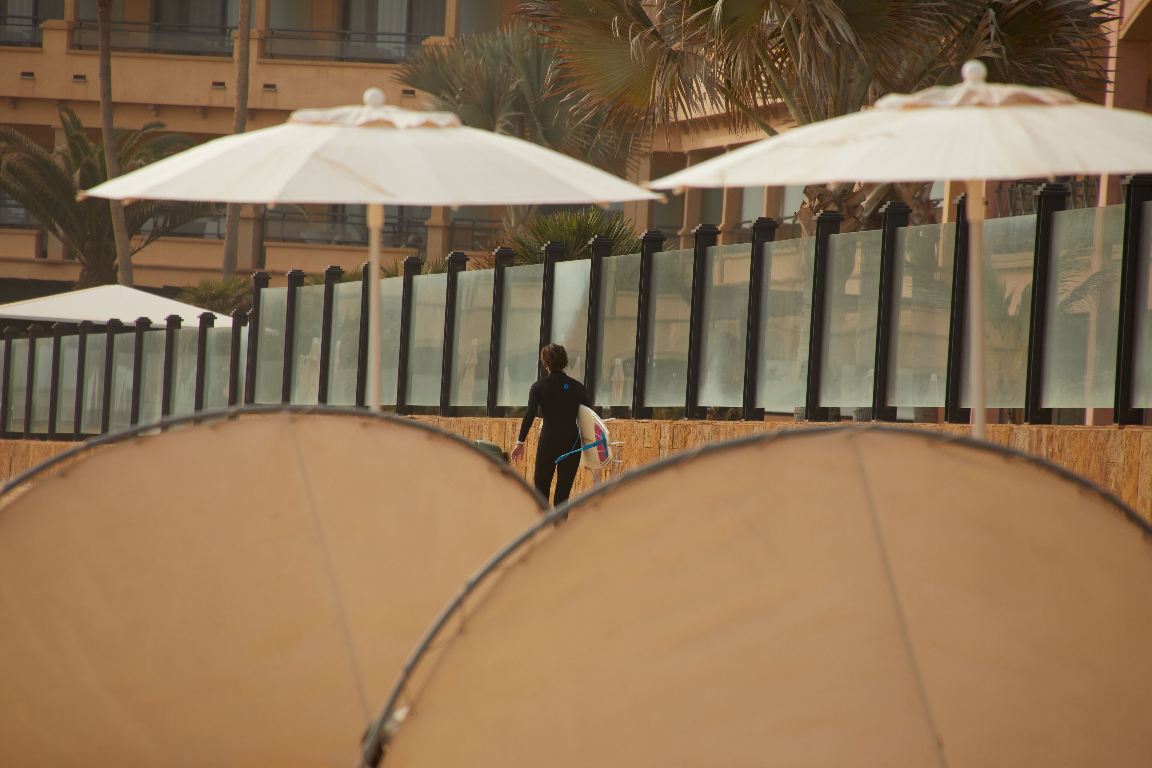 A person walking with a surfboard under umbrellas, palm trees, and a building in the background.