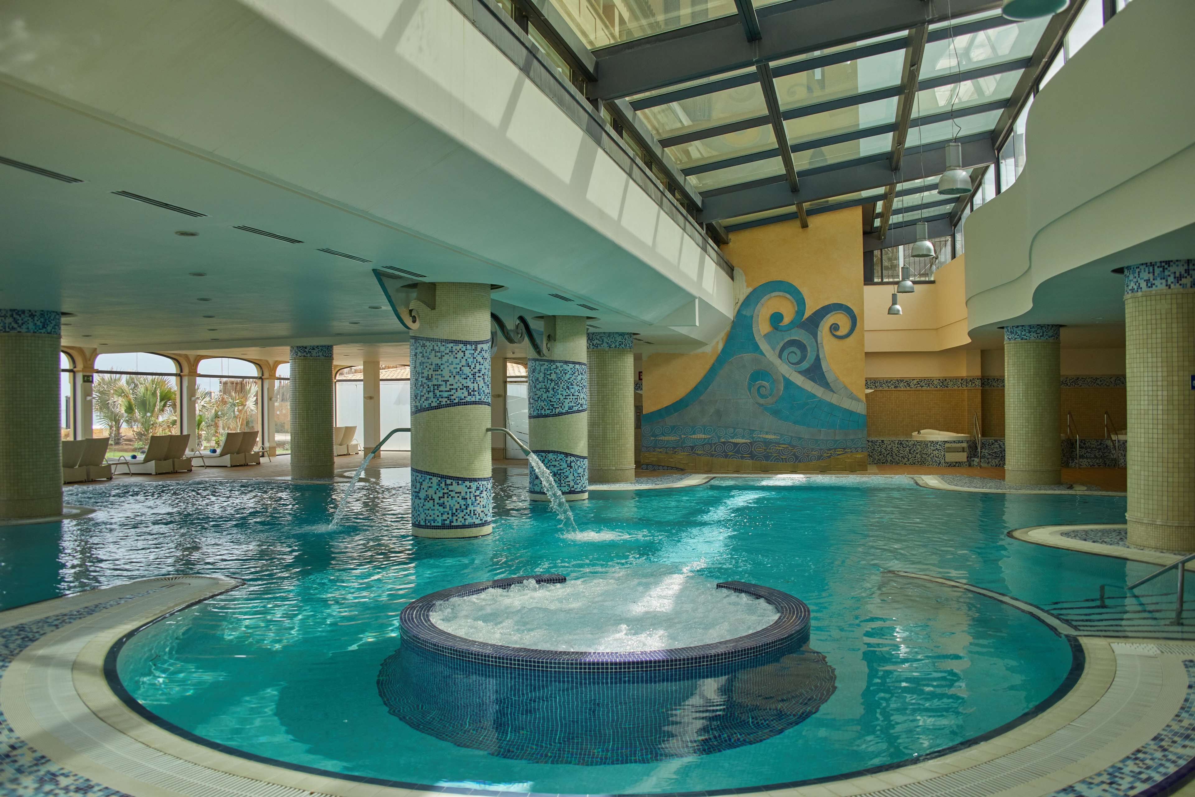 Una piscina interior con un jacuzzi central, paredes de azulejos y un techo alto con tragaluces.