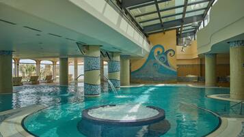 Una piscina interior con un jacuzzi central, paredes de azulejos y un techo alto con tragaluces.