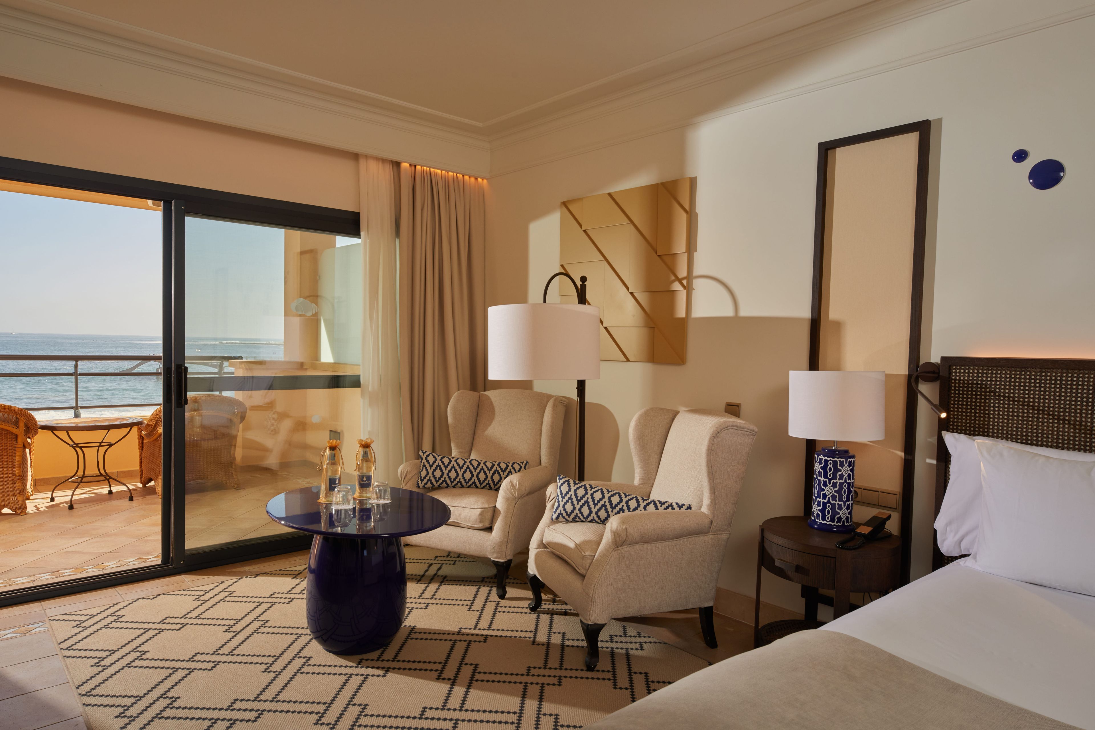 A hotel room with a large window, a round table, two armchairs, a bed, and a nightstand with a lamp.