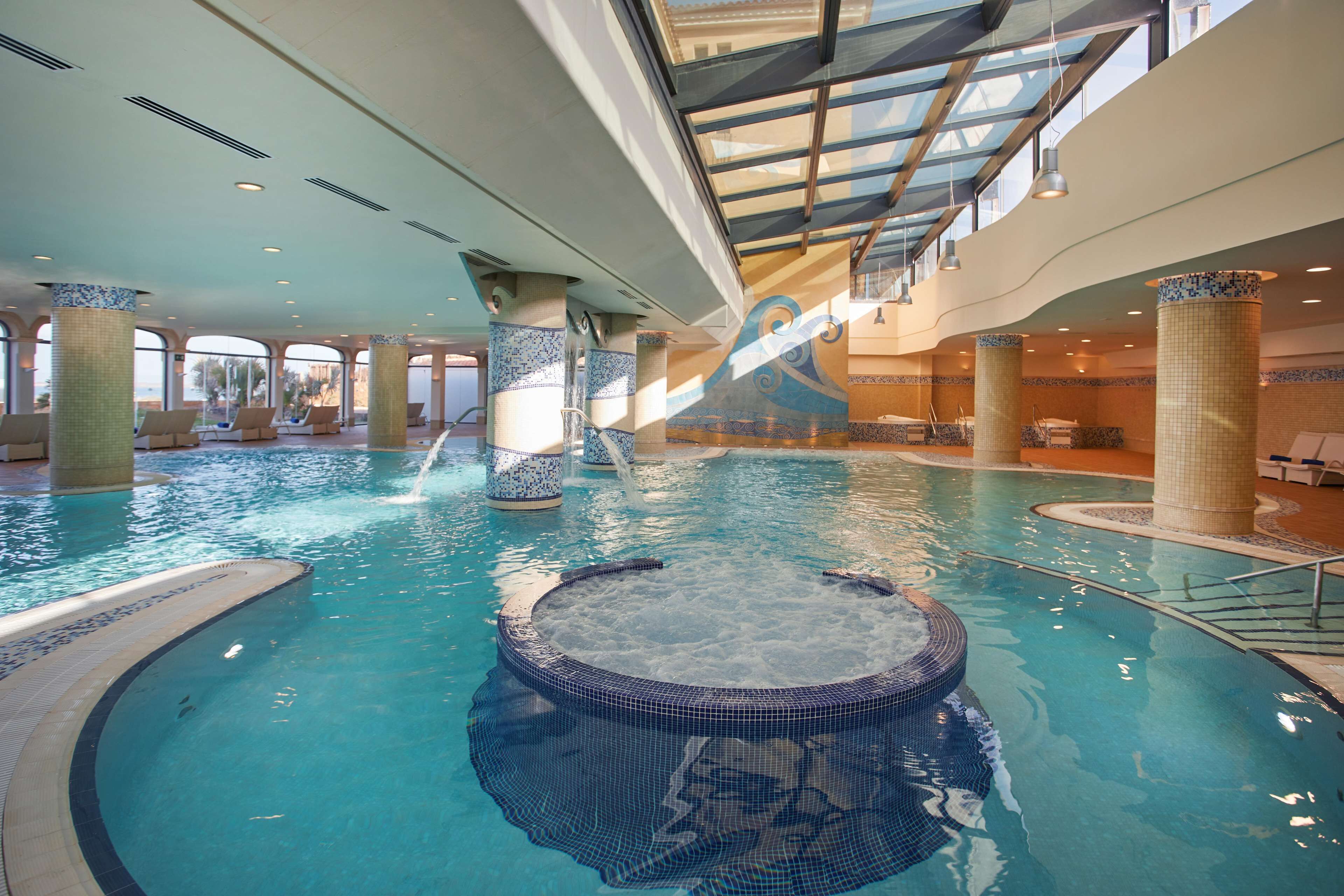 Uma piscina interior com hidromassagem, colunas revestidas de azulejos e teto de vidro.