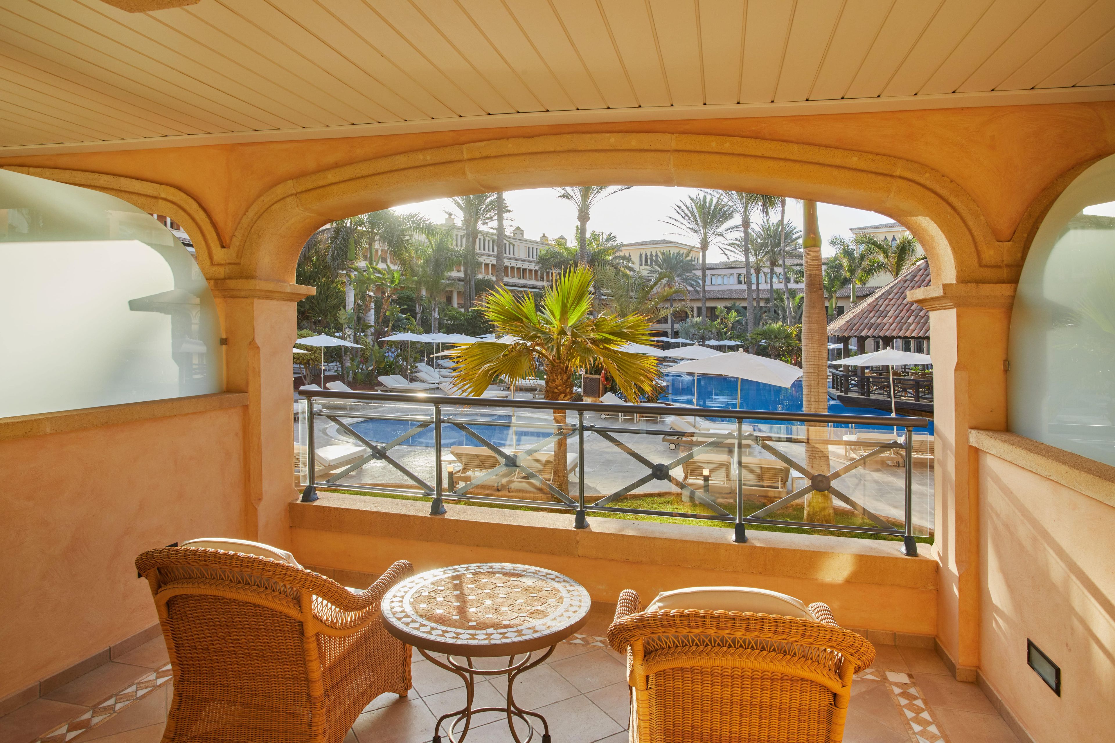 Un balcon doté de meubles en osier, d’une rambarde en verre et offrant une vue sur une piscine et des palmiers.