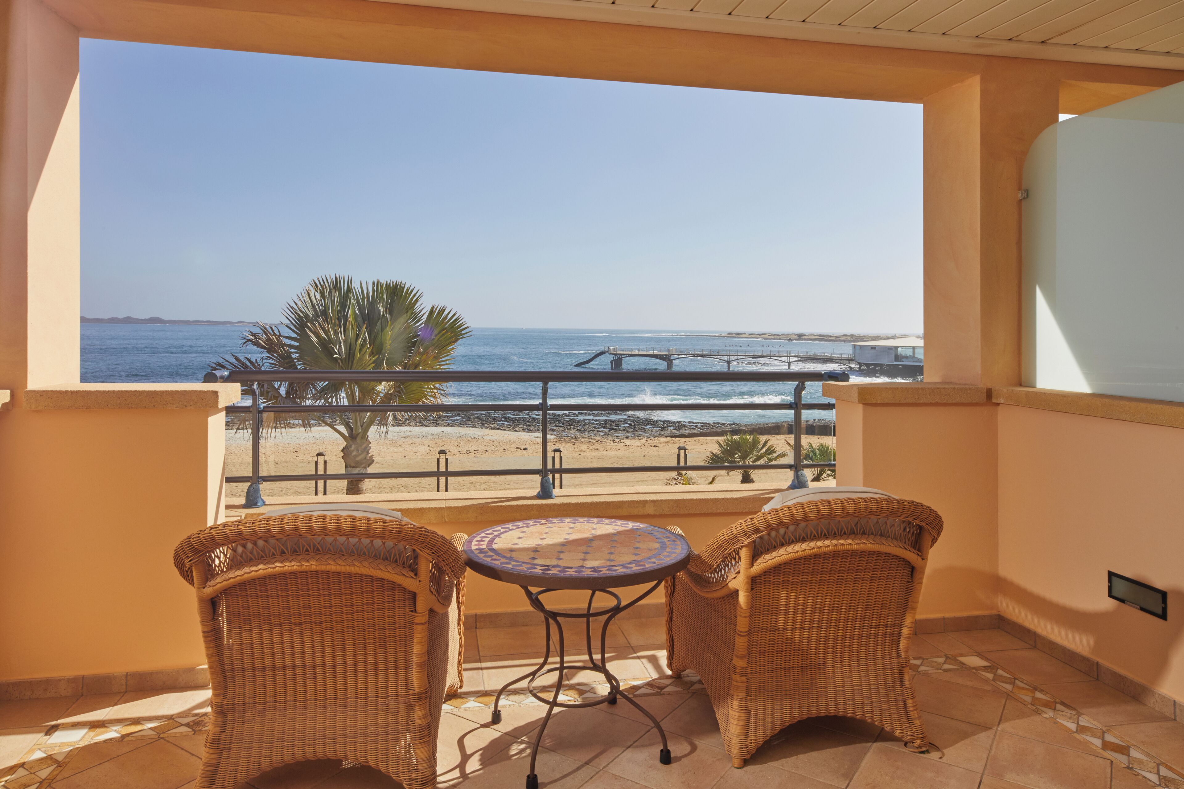 A balcony with wicker chairs and a small table, overlooking a beach and the sea.