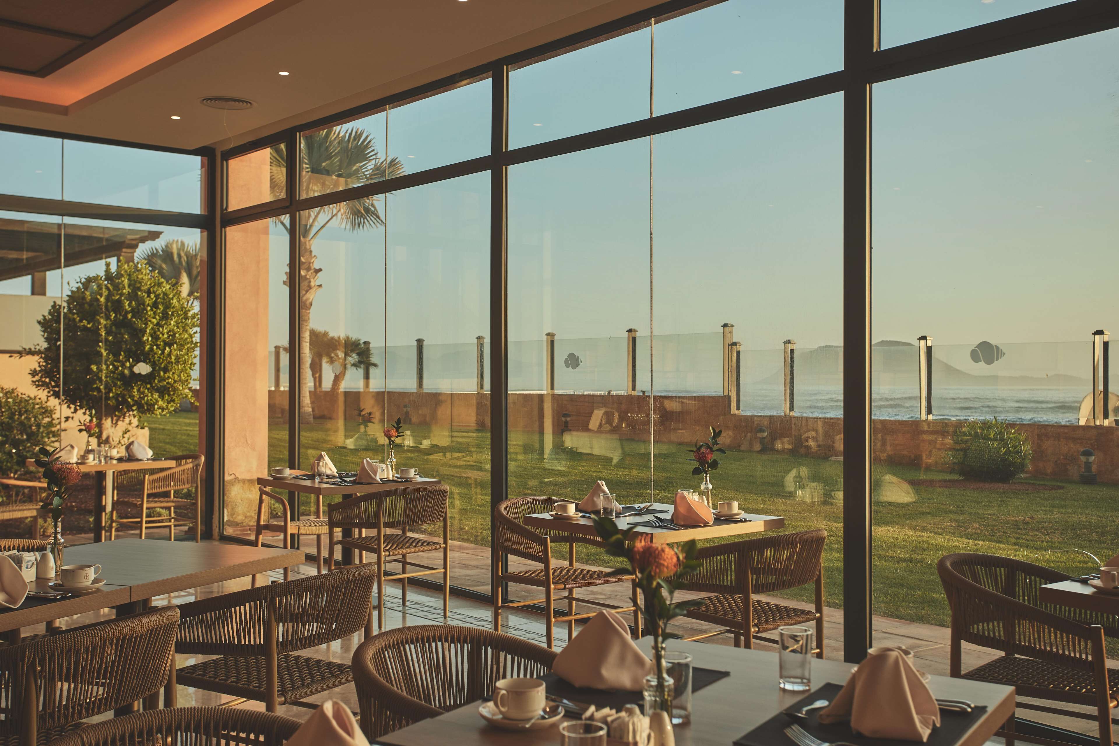 A restaurant with large windows offering a view of the ocean and a grassy area. Tables are set with chairs and dining ware.