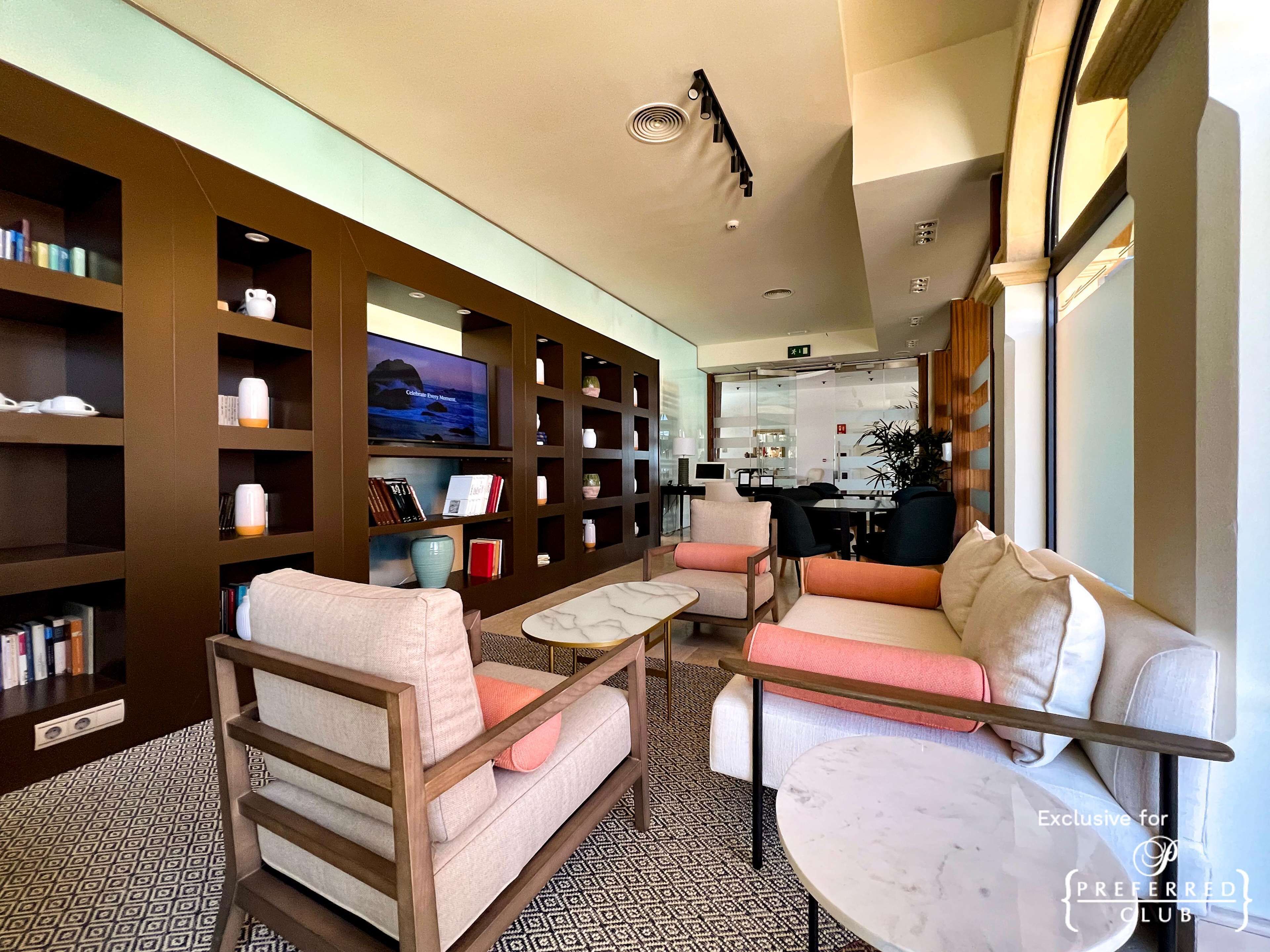 A modern living room with a sofa, armchairs, a coffee table, and a television. There are bookshelves with various items and a patterned carpet.