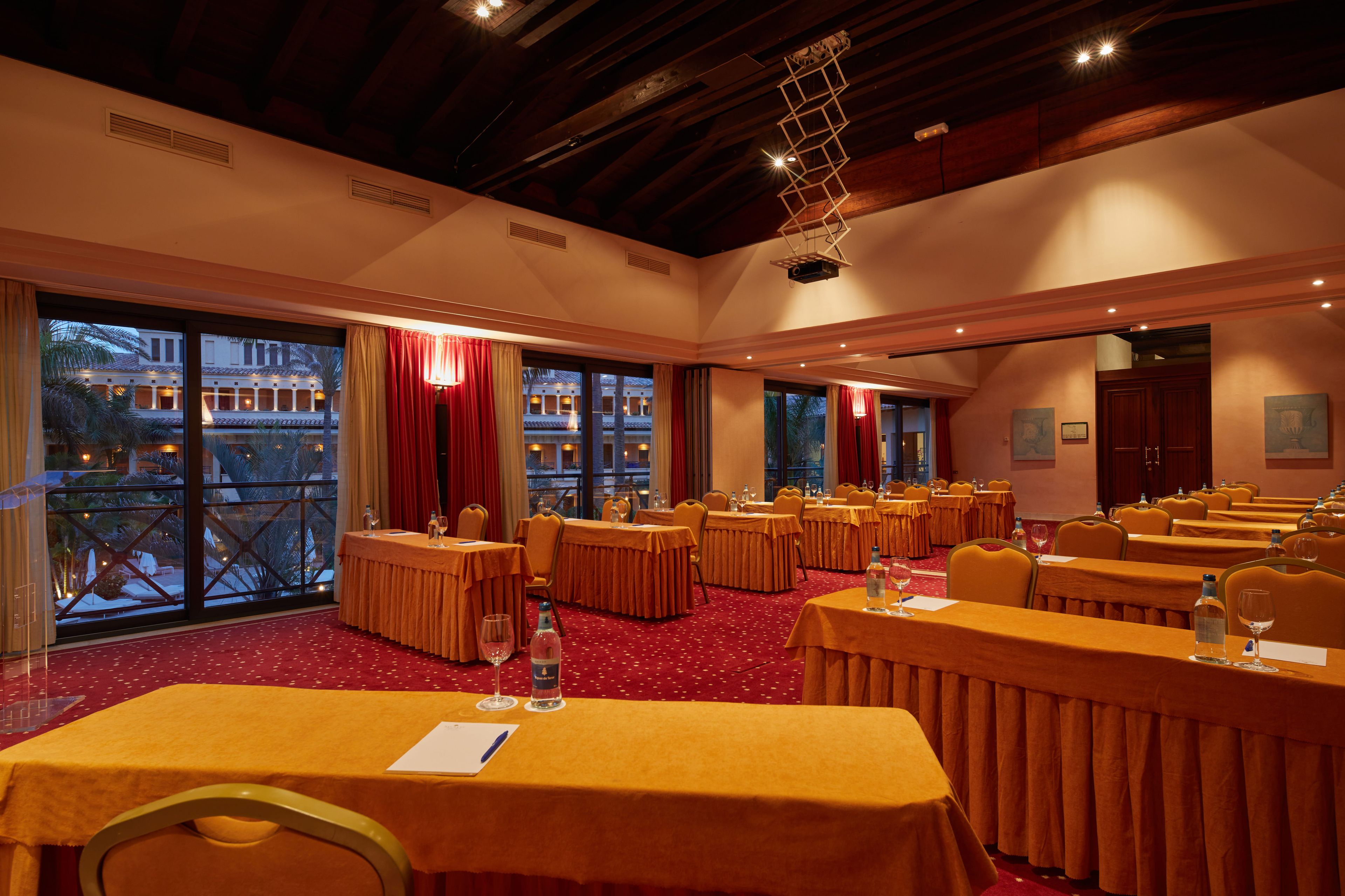 Un salón de conferencias con varias mesas, cortinas rojas y grandes ventanales con vista a un edificio.