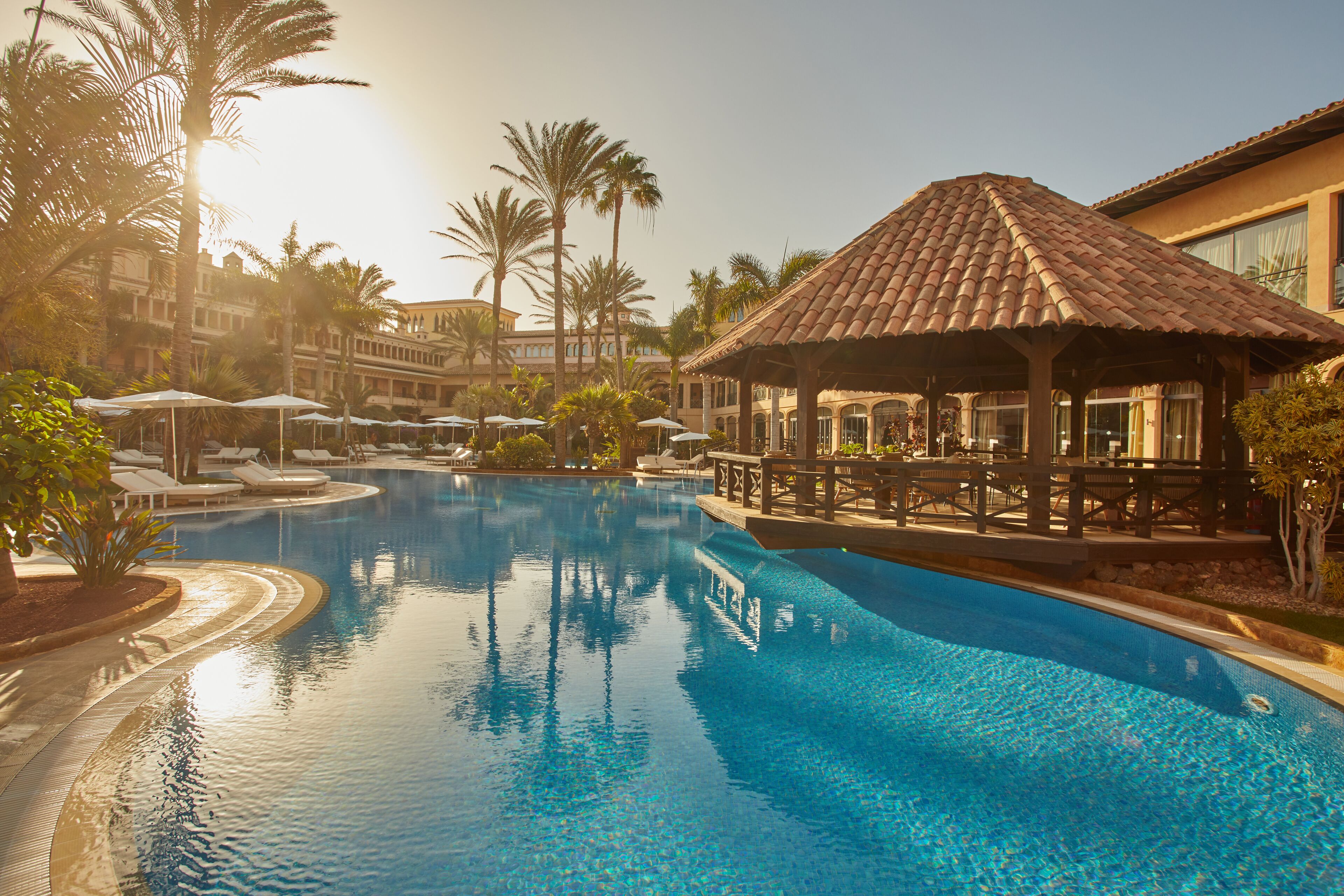 A swimming pool with lounge chairs and umbrellas, surrounded by palm trees and a building with a tiled roof.