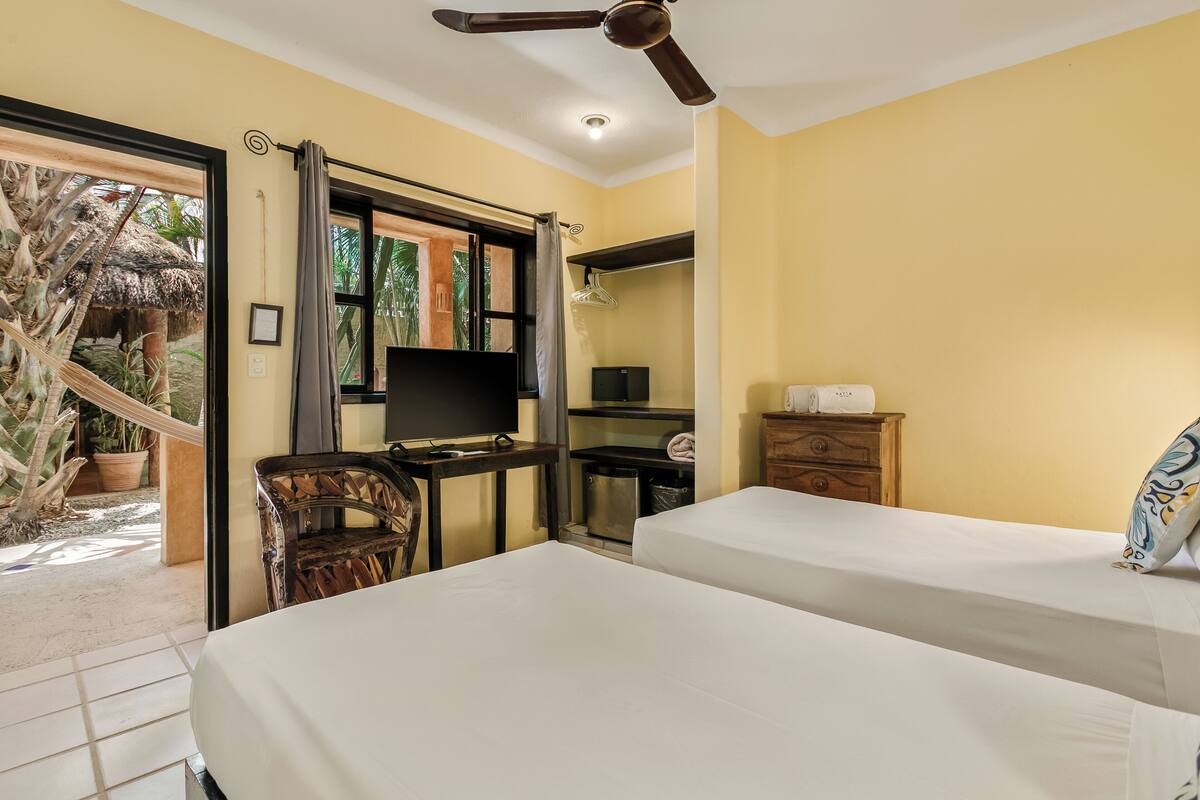 Une chambre avec deux lits, une télévision, une commode en bois, un bureau avec une chaise et un hamac à l’extérieur.