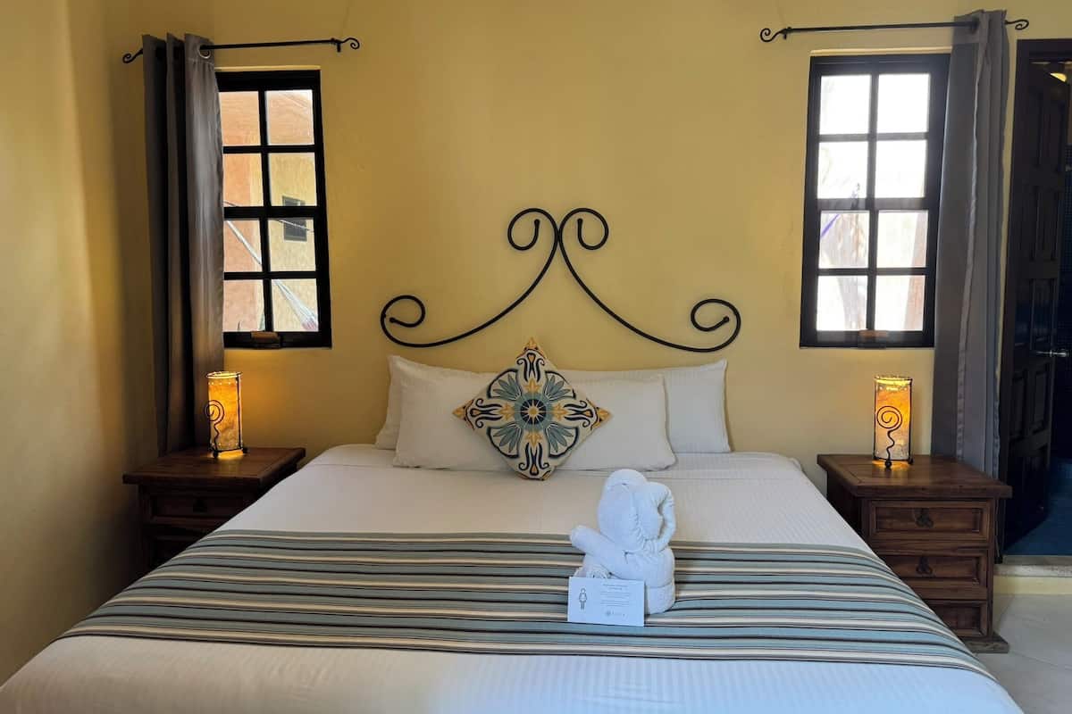 Un lit bien fait, avec une literie à rayures, un coussin décoratif et un ours en peluche posé sur le lit. Il y a deux tables de chevet avec des lampes, et la pièce présente un aménagement simple et épuré, avec un mur uni et une fenêtre agrémentée de rideaux.