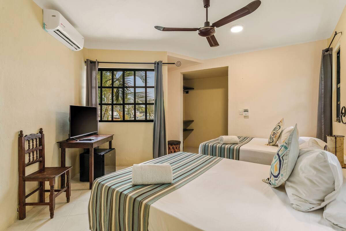 Une chambre d’hôtel avec deux lits, un bureau avec une télévision, un climatiseur, un ventilateur de plafond et une fenêtre avec des rideaux.