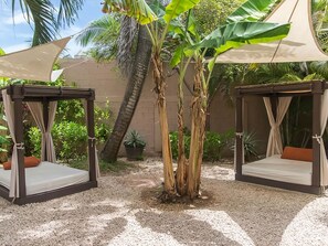 Dos camas de madera al aire libre con cojines color beige, rodeadas de grava, bajo grandes toldos, con una palmera y plantas en macetas al fondo.