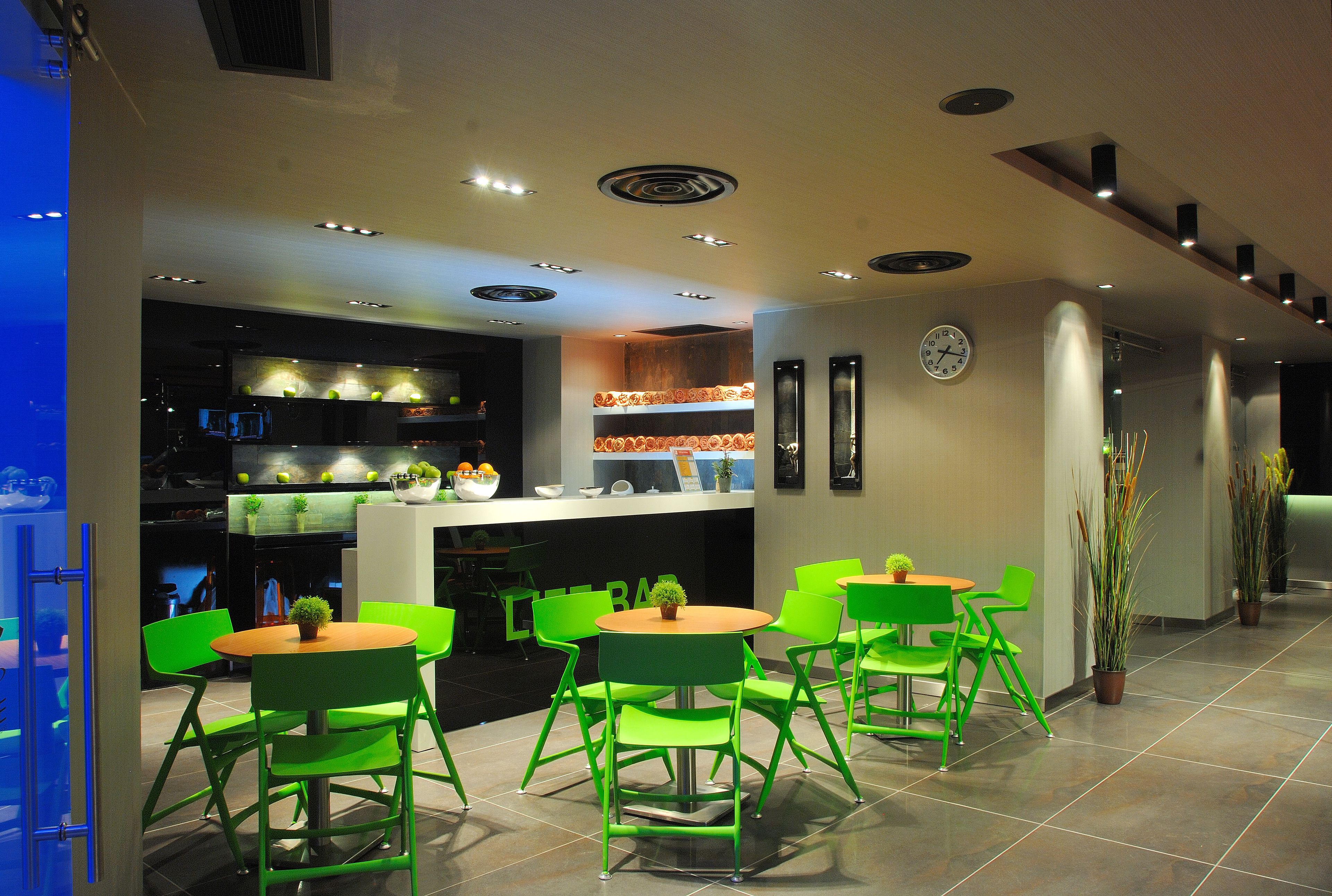A modern restaurant with green chairs and tables, a kitchen area, and potted plants.