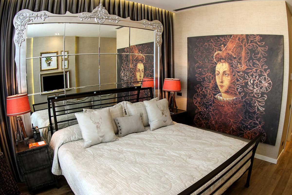 A bedroom with a large mirror, a bed with multiple pillows, two bedside lamps, and a floral wallpaper.