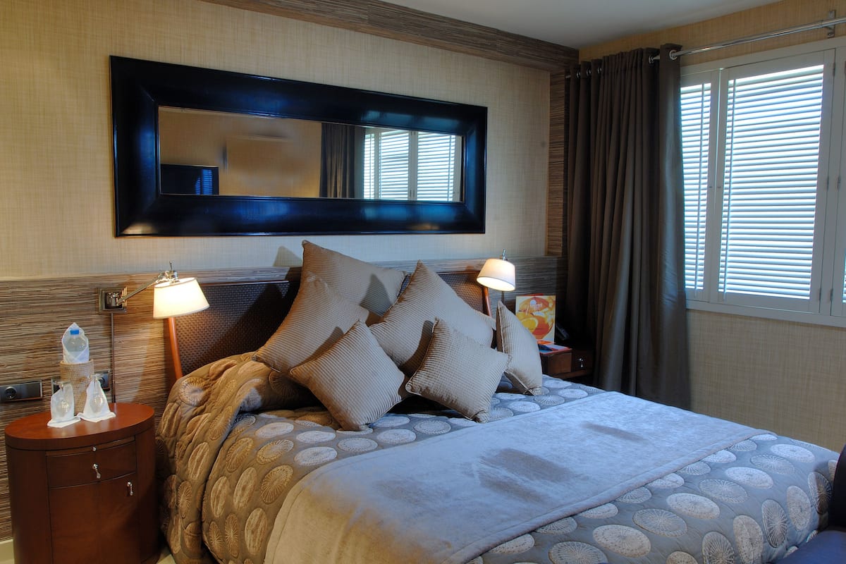 A hotel room with a large bed, a nightstand with a lamp, a mirror above the bed, and a window with curtains.