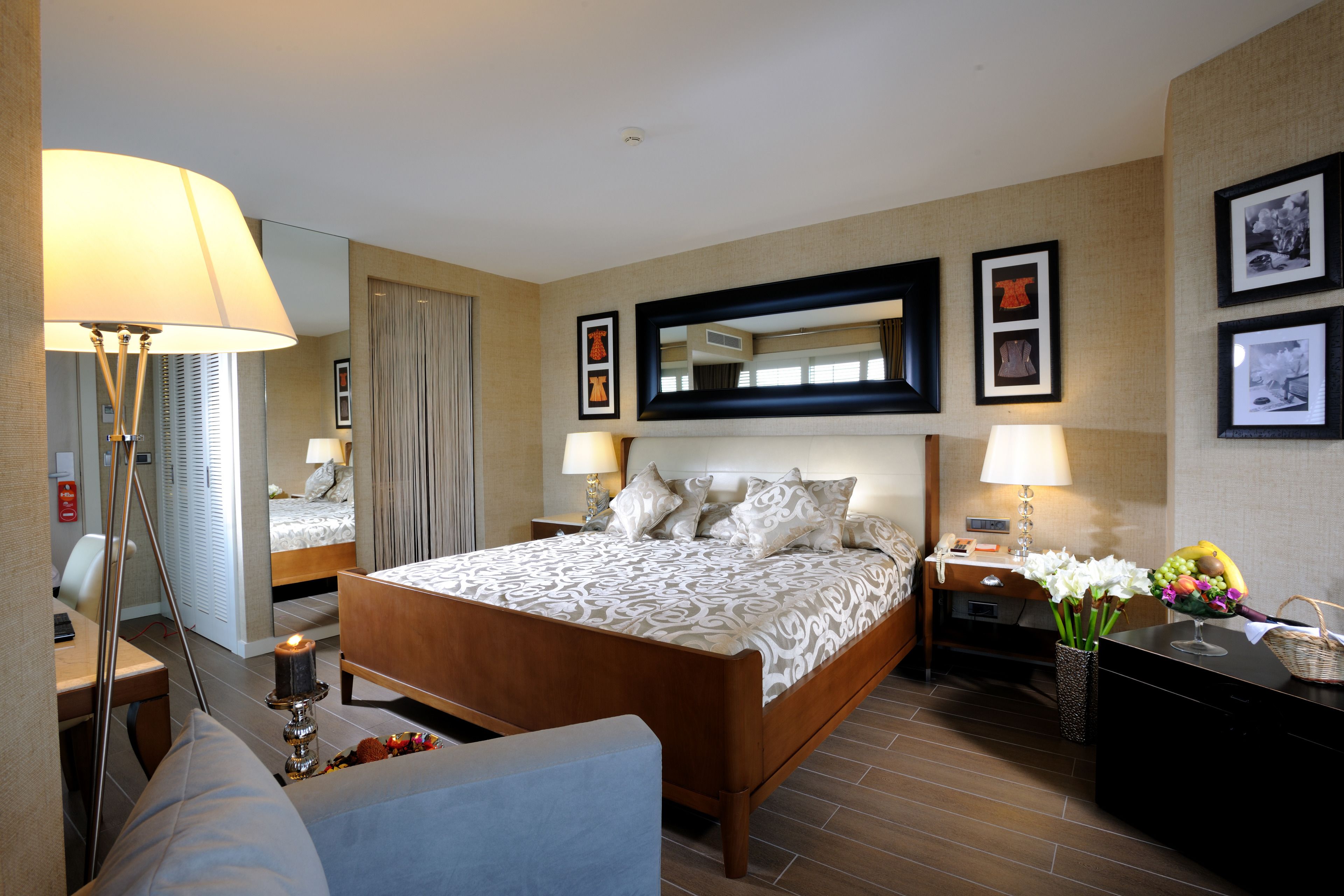 A hotel room with a bed, bedside tables, a desk, a chair, a lamp, a mirror, and framed pictures on the wall.