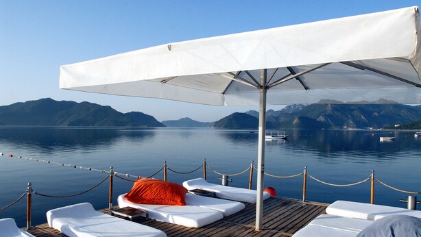 A deck with lounge chairs and an umbrella, overlooking calm water and mountains.