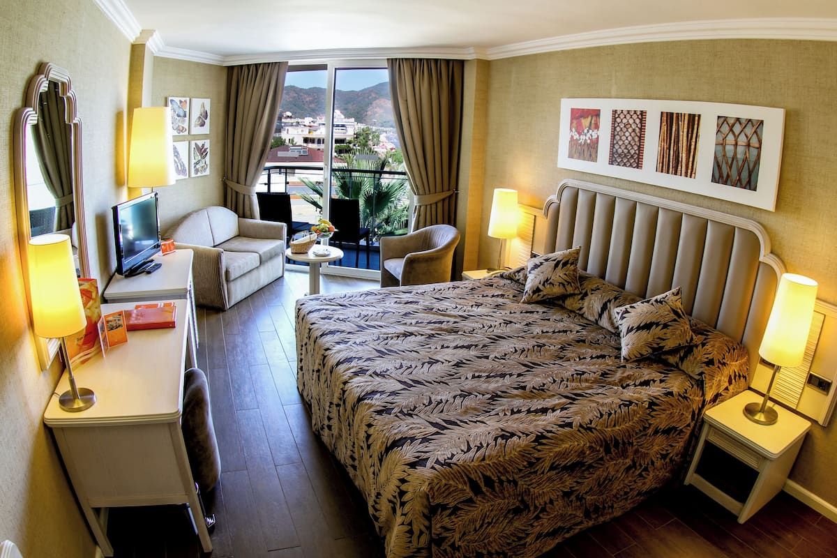 A hotel room with a bed, a desk, a chair, a small table, and a balcony with a view of the city.