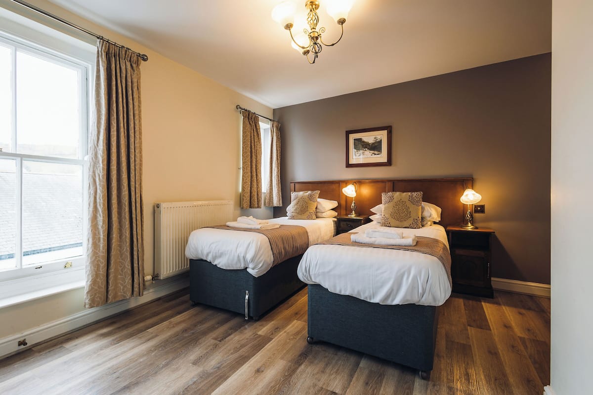 Une chambre d’hôtel avec deux lits, un radiateur, une fenêtre avec des rideaux, un lustre et un tableau encadré au mur.
