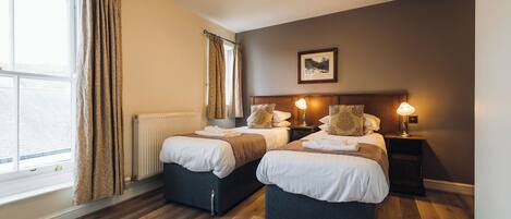 A hotel room with two beds, a radiator, a window with curtains, a chandelier, and a framed picture on the wall.