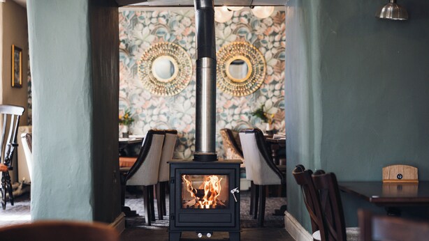A cozy room with a wood-burning stove, patterned wallpaper, and a dining area with chairs and a table. - White Lion Royal Hotel (Bala)