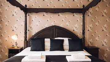 A four-poster bed with a dark wooden frame, white bedding, and black pillows. A small bedside table with a lamp is visible to the left. The wall features a repeating pattern of white birds.