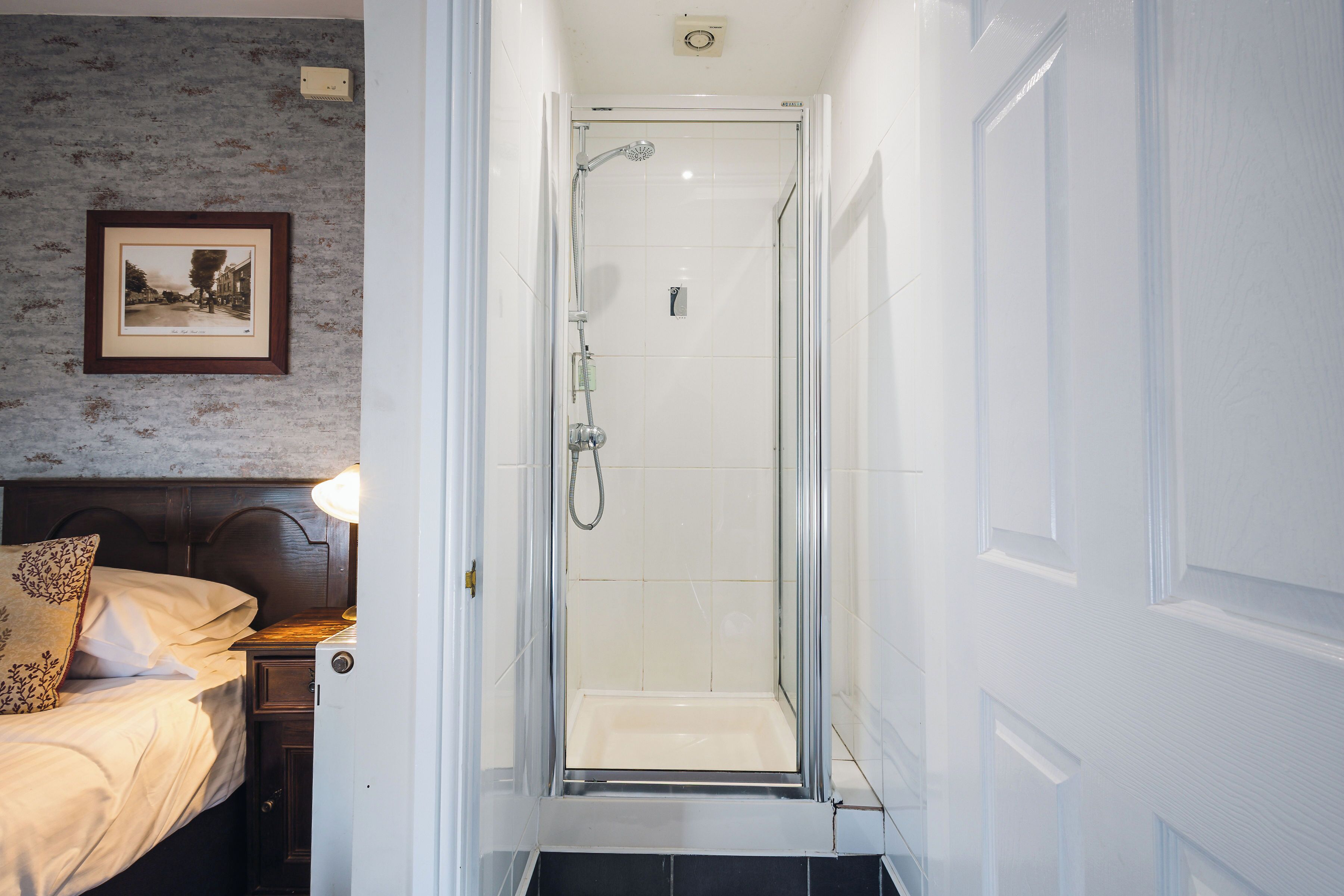 A narrow bathroom with a shower, a bed with a headboard, and a framed picture on the wall.