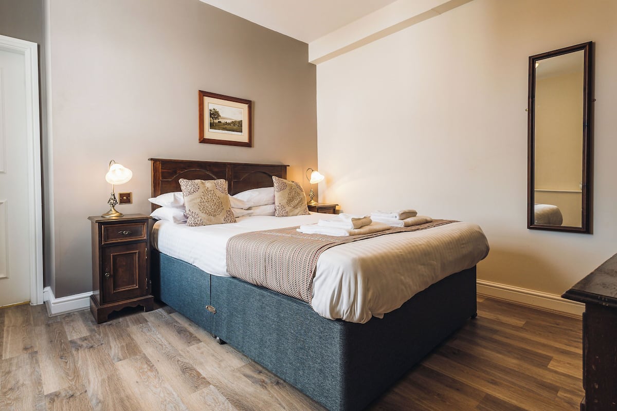 Un lit bien fait, avec une tête de lit, deux tables de chevet avec des lampes, un tableau encadré au mur et un grand miroir.