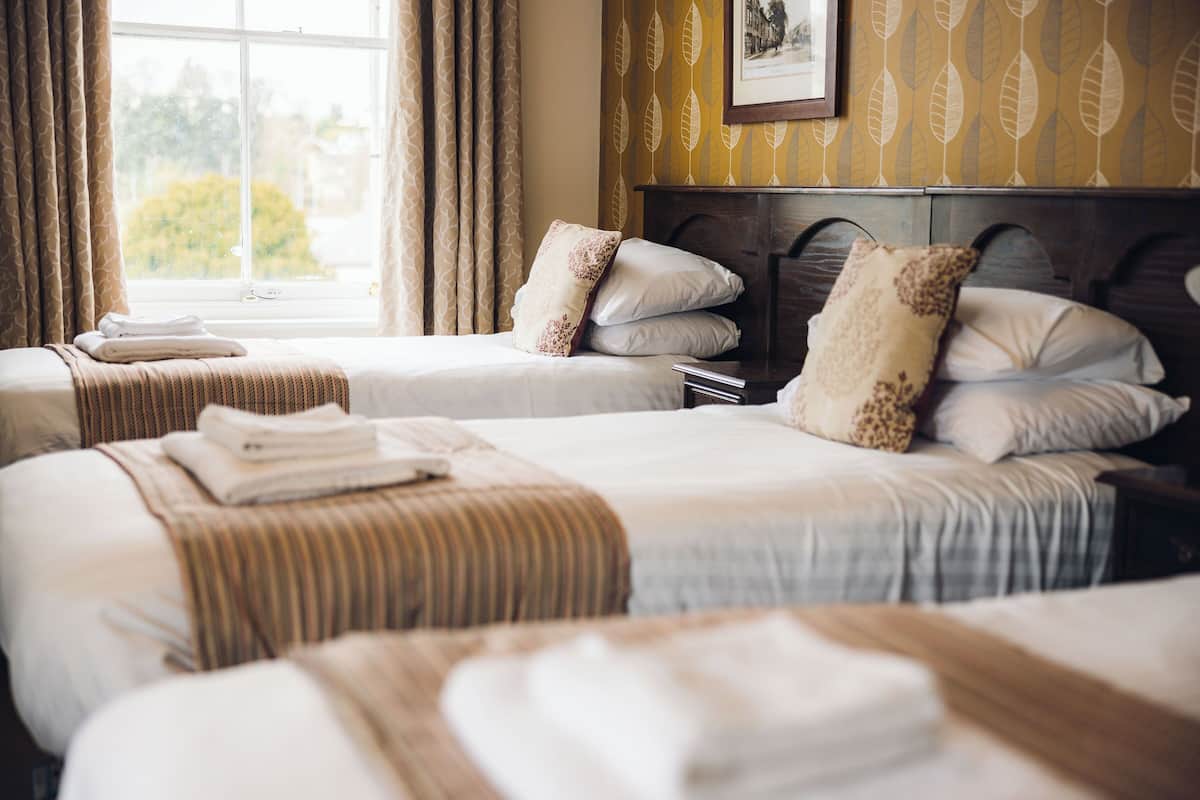 Une chambre d’hôtel avec deux lits, chacun équipé d’un oreiller et d’une serviette pliée. La chambre dispose d’une fenêtre avec des rideaux, d’une tête de lit en bois et d’un mur orné d’un motif de feuilles.