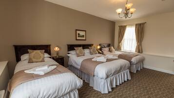 A hotel room with three beds, each with white linens and decorative pillows. There is a chandelier, a framed picture on the wall, and a window with curtains.