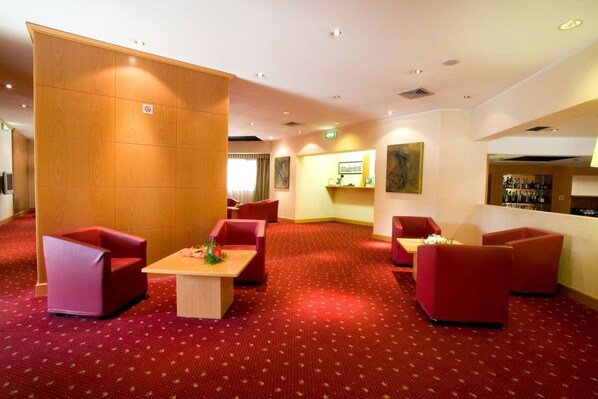 Um saguão de hotel com piso em carpete vermelho, assentos vermelhos, uma divisória de madeira e uma área de bar com garrafas.