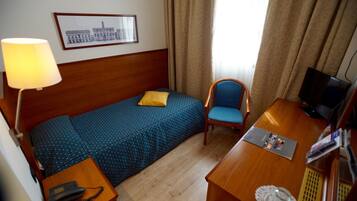A hotel room with a bed, a desk, a chair, a lamp, a TV, and a framed picture on the wall.