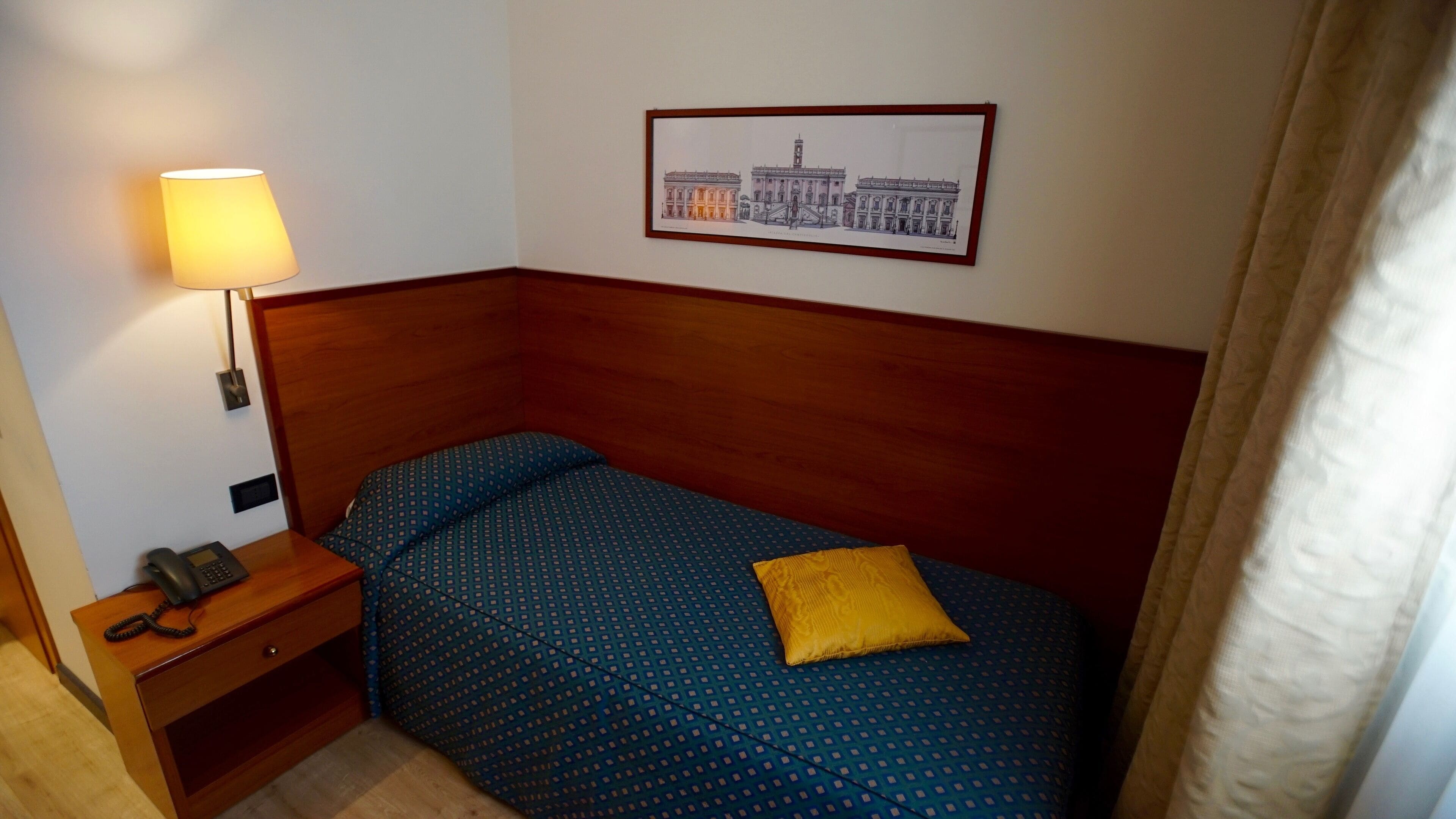 Una camera da letto piccola e ordinata con un letto singolo, un comodino con una lampada, un telefono e un quadro appeso al muro.