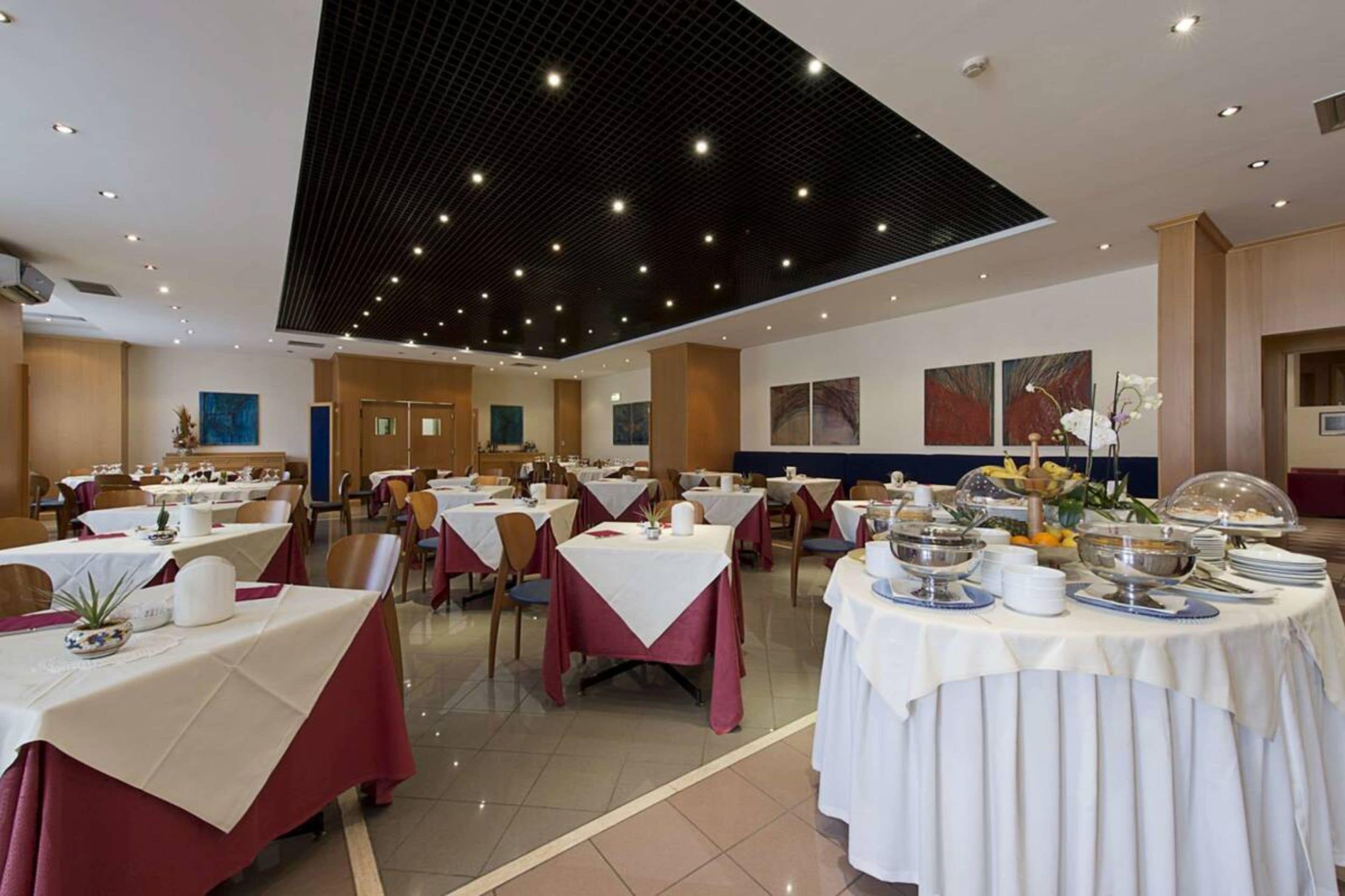 Un comedor amplio con varias mesas dispuestas para el desayuno, que destaca por su moderno diseño de techo y obras de arte en las paredes.