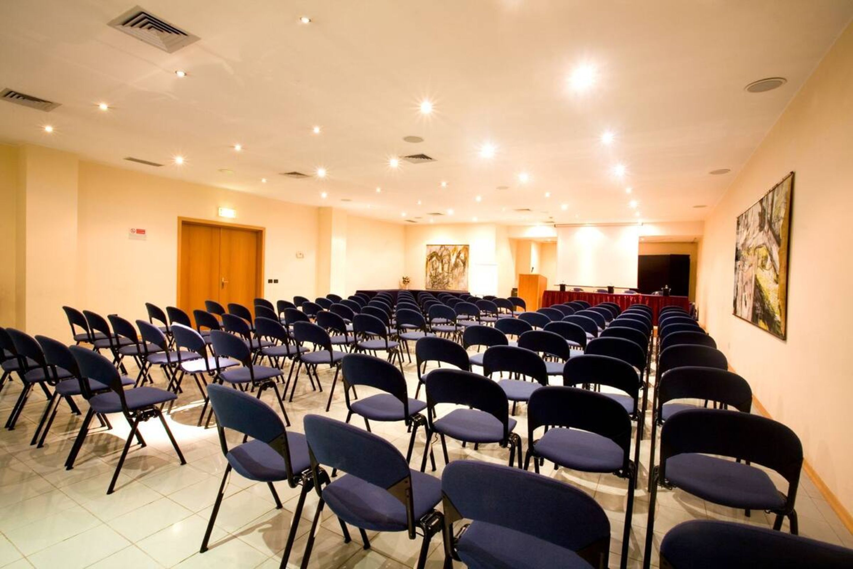 Un salón de conferencias con filas de sillas, un atril y un cuadro grande en la pared.