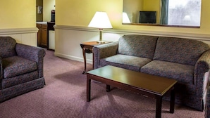 A living room with a sofa, armchair, and a small table. There is a lamp on a side table, a ceiling fan, and a window with a view of trees. - Rodeway Inn & Suites Plymouth Hwy 64 (Plymouth)