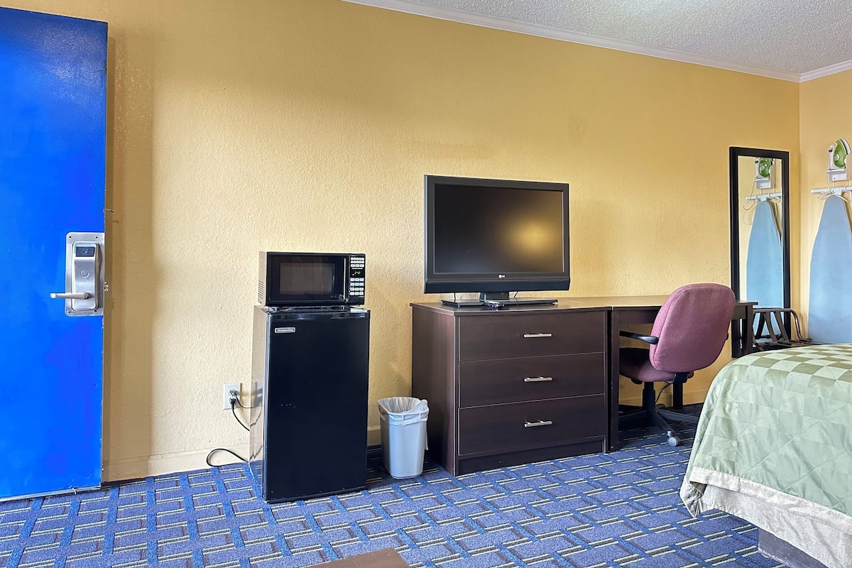 Ein Hotelzimmer mit einem Bett, einem Schreibtisch mit Computer und Fernseher, einem Sessel, einem Mülleimer und einer Tür.