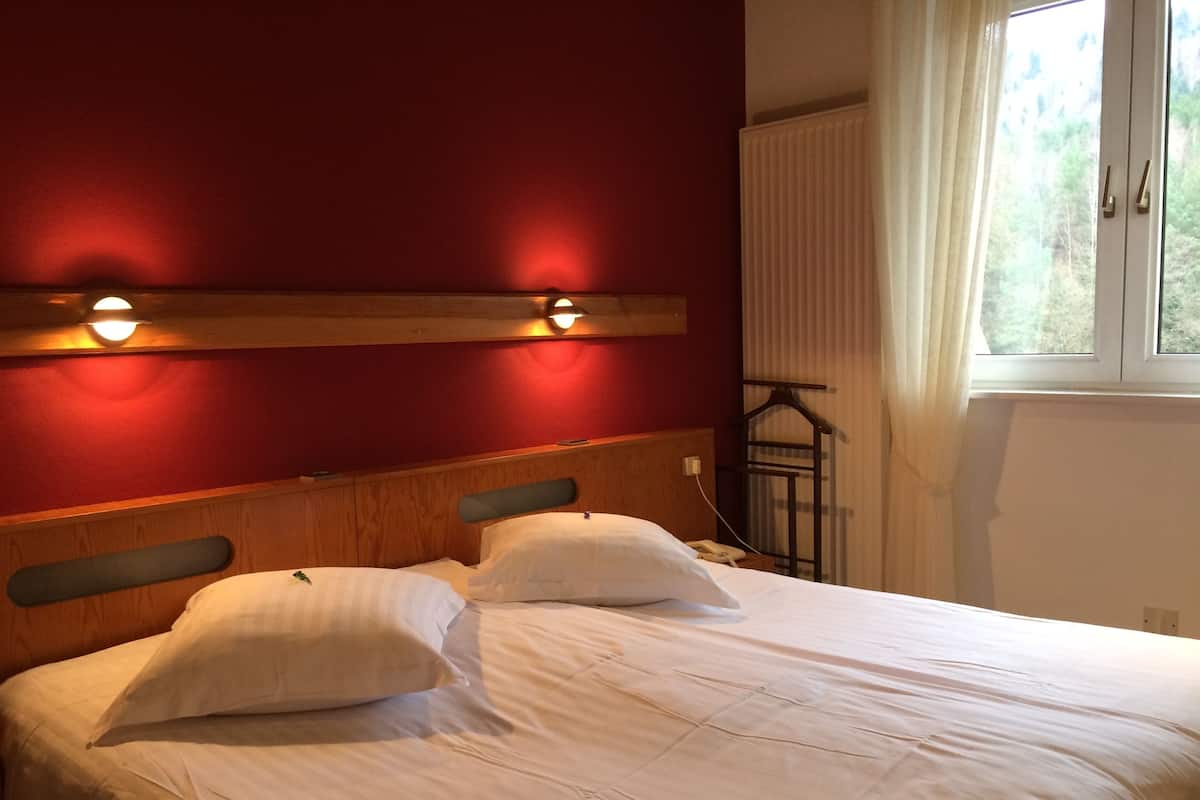 Un lit avec du linge de lit blanc et deux oreillers, une tête de lit en bois agrémentée de deux lampes murales, et une fenêtre donnant sur un paysage verdoyant.