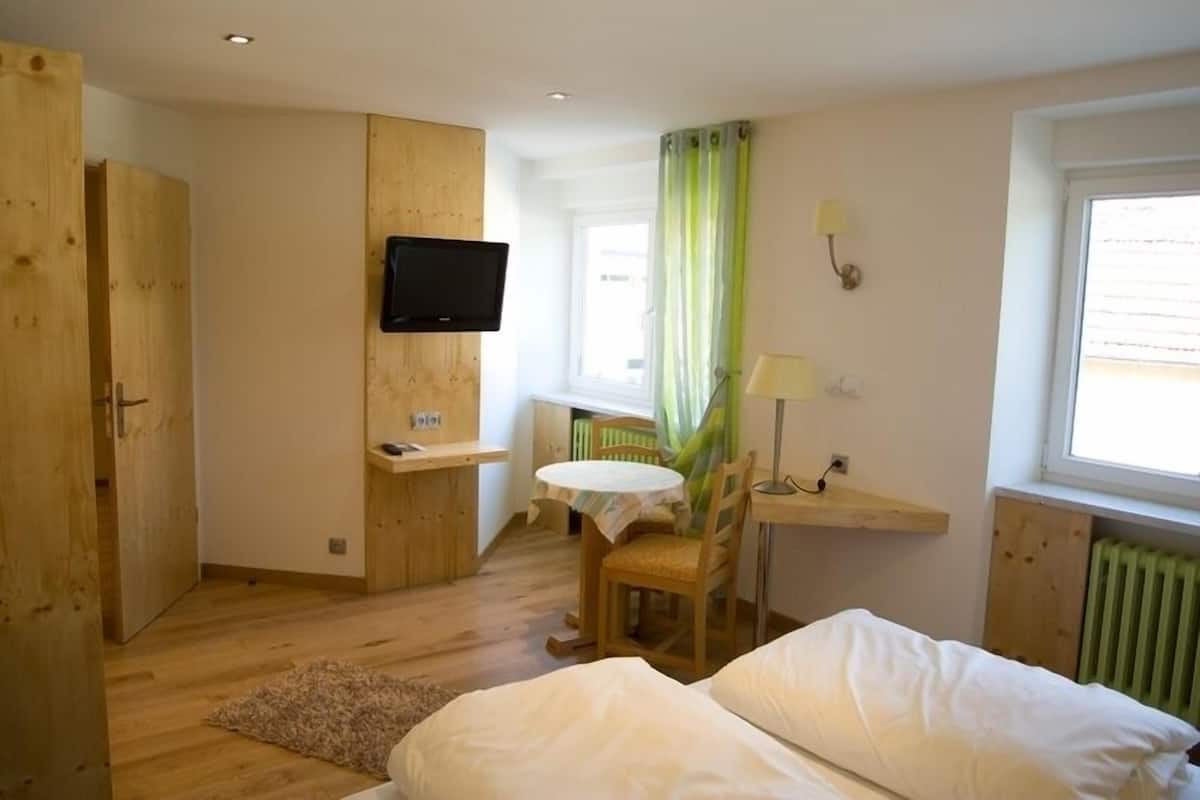 Une petite pièce comprenant un lit, un bureau, une chaise, un téléviseur et une fenêtre avec des rideaux.