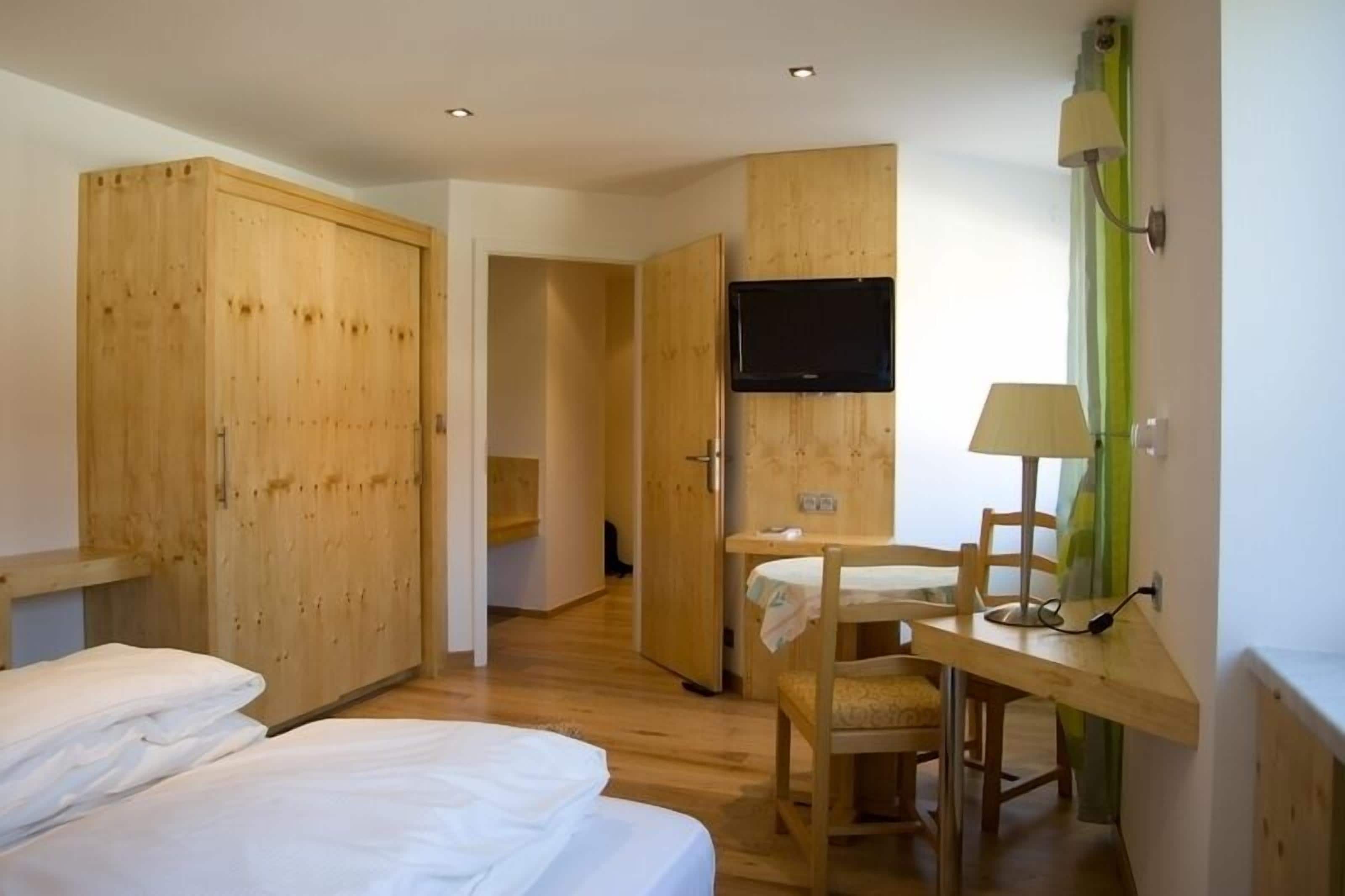 A hotel room with wooden wardrobes, a bed, a desk with a chair, a television, and a lamp.