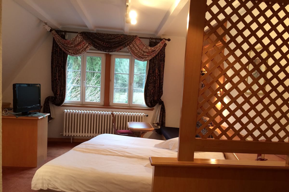 Une chambre à coucher avec un lit, un bureau avec un ordinateur, une fenêtre avec des rideaux et un radiateur.
