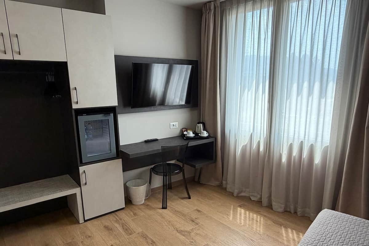 Una habitación de hotel moderna con kitchenette, un televisor de pantalla plana, un escritorio con silla, un pequeño cesto de basura y una ventana con cortinas.