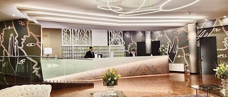 A modern hotel lobby with a reception desk, a glass partition, a white sofa, a glass coffee table, and a patterned carpet.