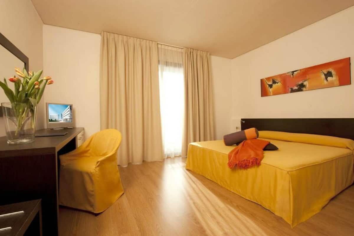 Habitación de hotel con cama, escritorio, silla, un cuadro en la pared y ventana con cortinas.